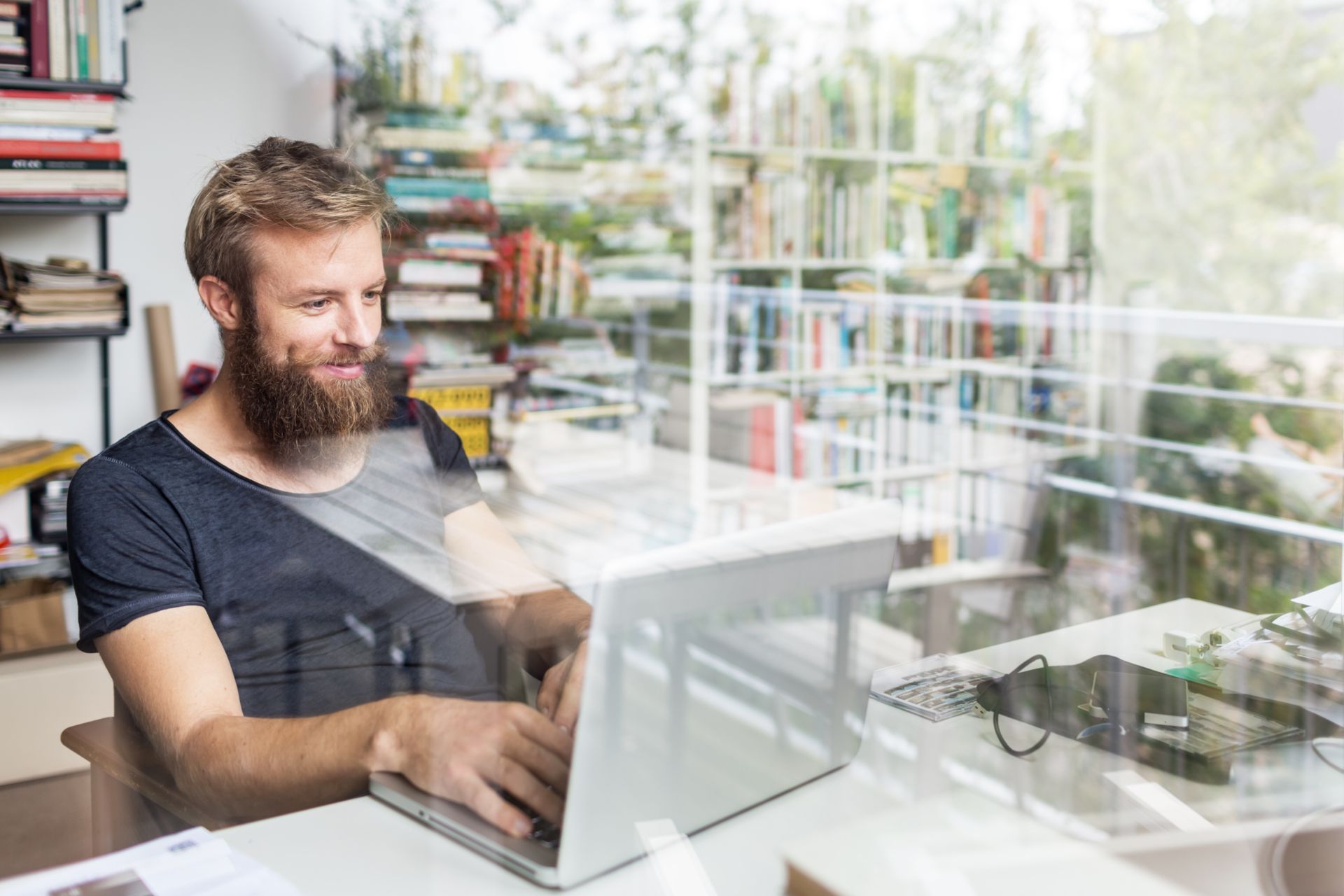 A man with a beard sits at a desk and uses a laptop in the context of CANCOM Austria's IT services.